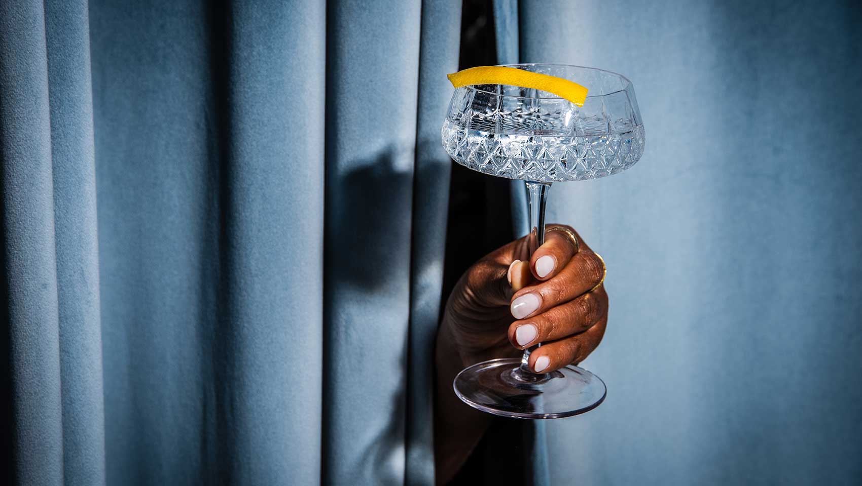 Hand peeking out from behind a curtain and holding a cocktail with a lemon rind garnish