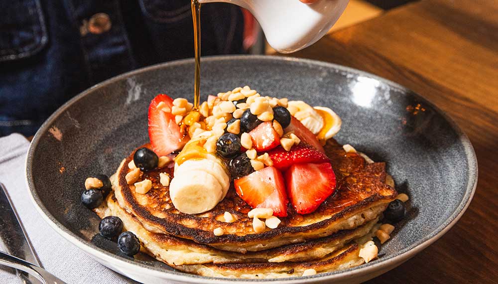 woman pouring syrup on a stack of pancakes with fruit on top at Blue Hound Kitchen